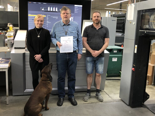 Sabine Krüger (Verantwortliche für die Vorstufe), Thomas Löhnert und Harald Weser (Drucker) (v. l. n. r.) freuen sich zusammen mit der "guten Seele" des Hauses, Firmenhund Gustav über die bestandene PSO-Zertifizierung